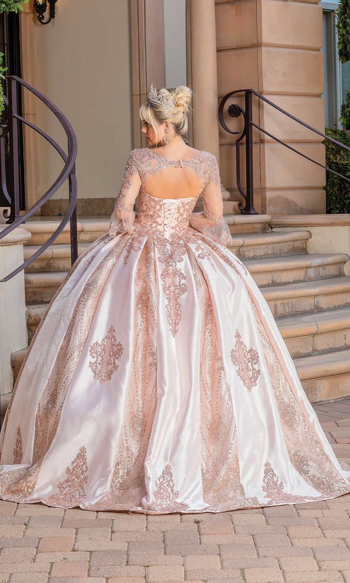 A woman in the Quinceanera Dress 1665 by Dancing Queen—an elaborate pink ball gown with lace details, sheer bell sleeves, and a tiara—stands at the base of stone steps facing away from the camera.